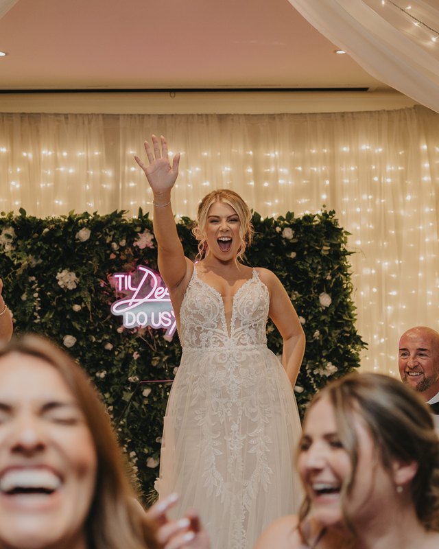A bride with her arm in the air on a packed dance floor as guests laugh and dance around her