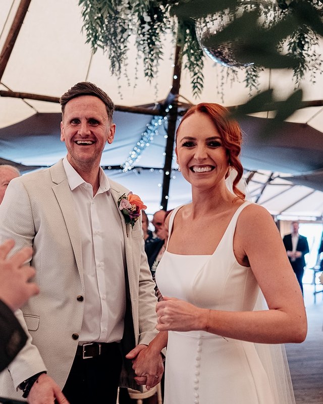 A beaming bride and groom holding hands at the front of their tipi ceremony as guests clap
