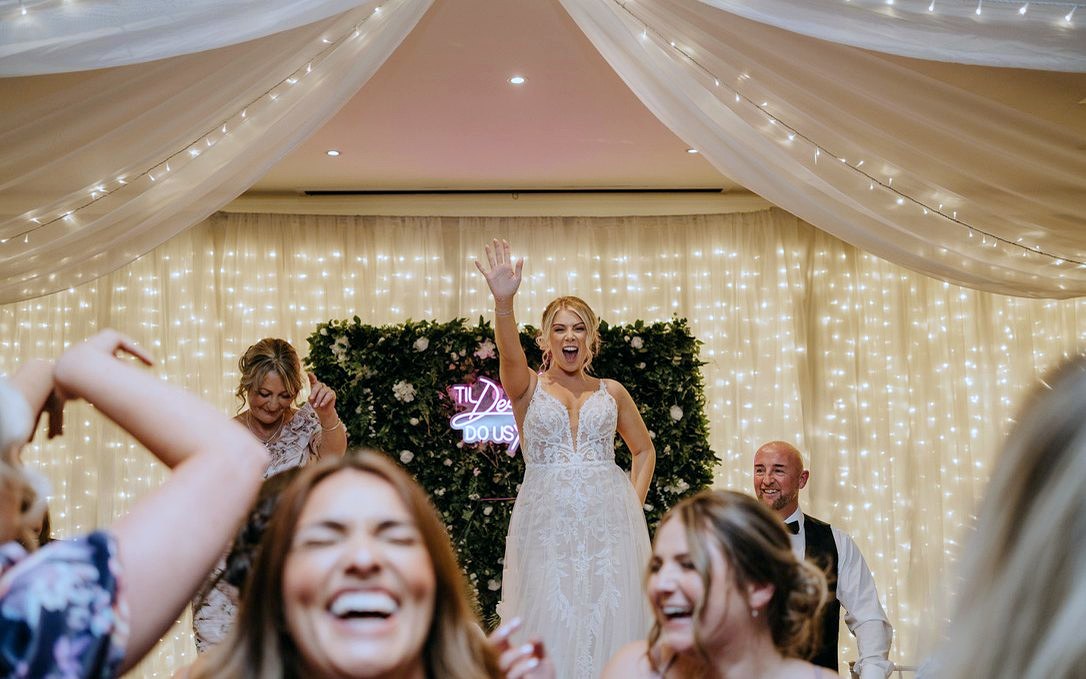 A bride standing on a chair with her arm in the air as guests laugh around her