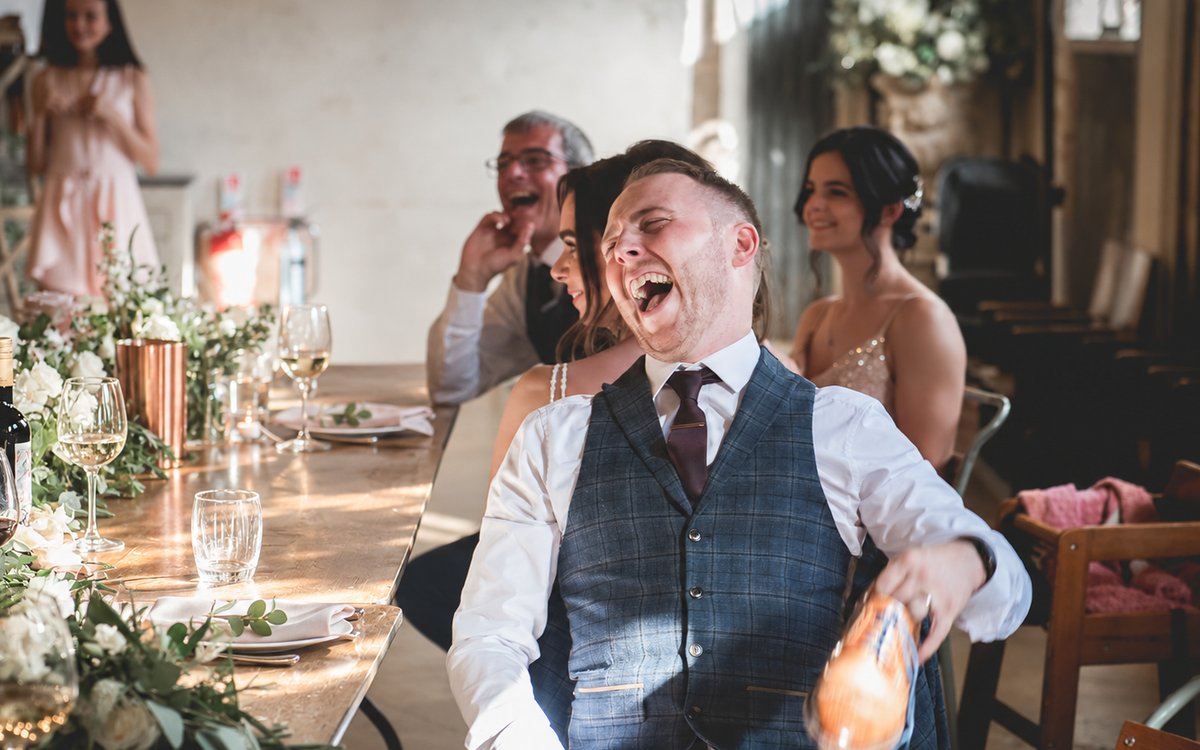 A groom crying with laughter at the top table, holding a shoe for the hot seat game