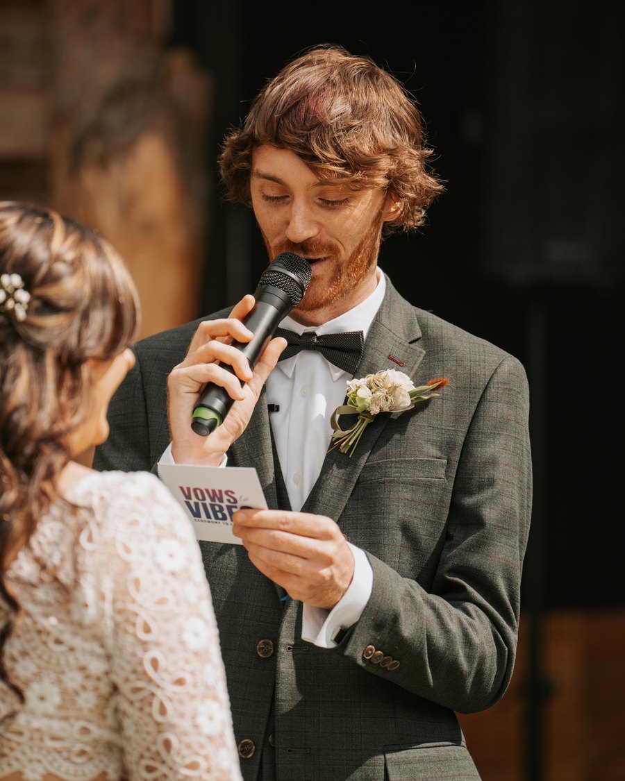 A groom reading his vows into a microphone from a Vows to Vibes card as his bride looks on